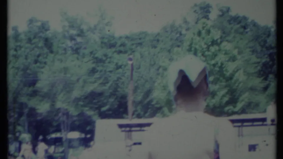 A person seen from behind wears a white hat near a low structure, with leafy trees filling the background in the 1971 outdoor scene.