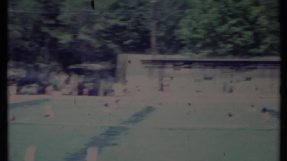 1971 California 8mm frame showing a sunlit swimming pool, grassy yard, trees, and a low residential structure in soft focus.