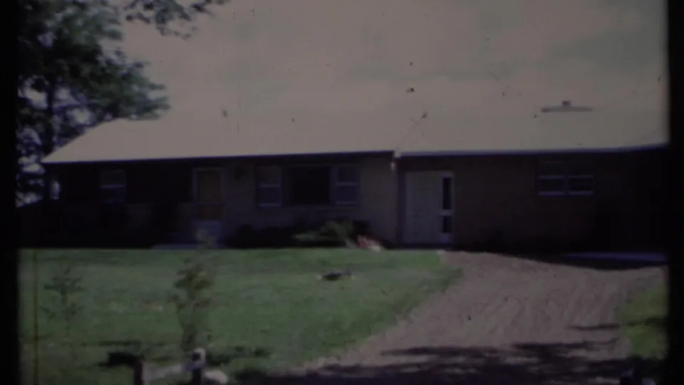 1971 California 8mm frame showing the house exterior, curved driveway, lawn, shrubs, and tree shade at the end of the pan.