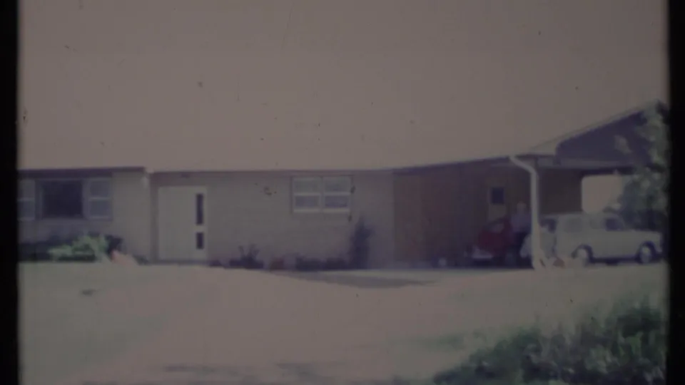 1971 California home-movie frame showing a house, carport, red car, white car, and a person near the vehicles.