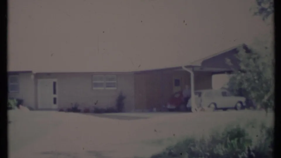 Faded 8mm frame of a California suburban home with a carport, red car, white car, driveway, and lawn.