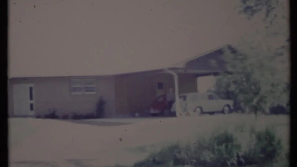 Soft 8mm view of a California suburban ranch house in 1971, with a shaded carport, two parked cars, shrubs, and a person near the vehicles.