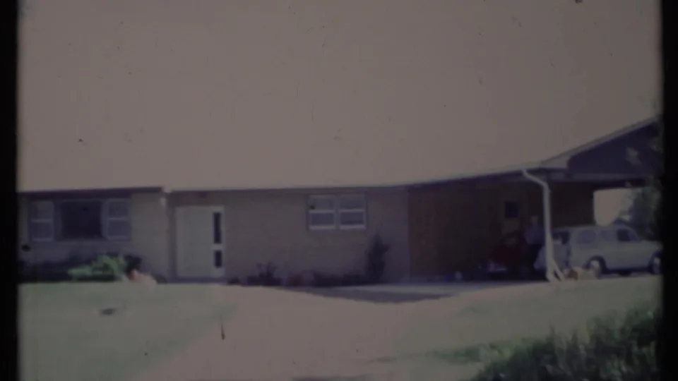 1971 California home-movie frame of a single-story house, showing the front door, broad window, driveway area, and cars partly visible under the carport.