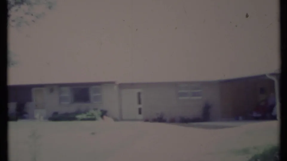 Faded 8mm frame of a 1971 California ranch house exterior, with lawn in front, large window, white door, shrubs, and a car partly visible at the carport.