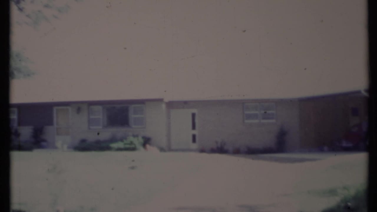 Light-colored single-story house with a white door, windows, driveway, lawn, and trees.