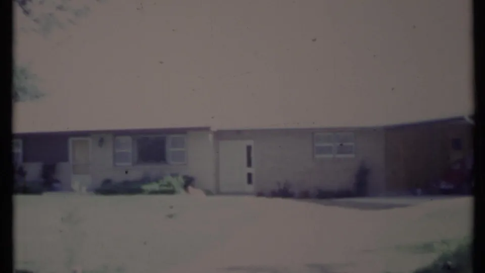 1971 California 8mm frame showing a light-colored suburban house exterior with a white door, windows, driveway, lawn, and trees.