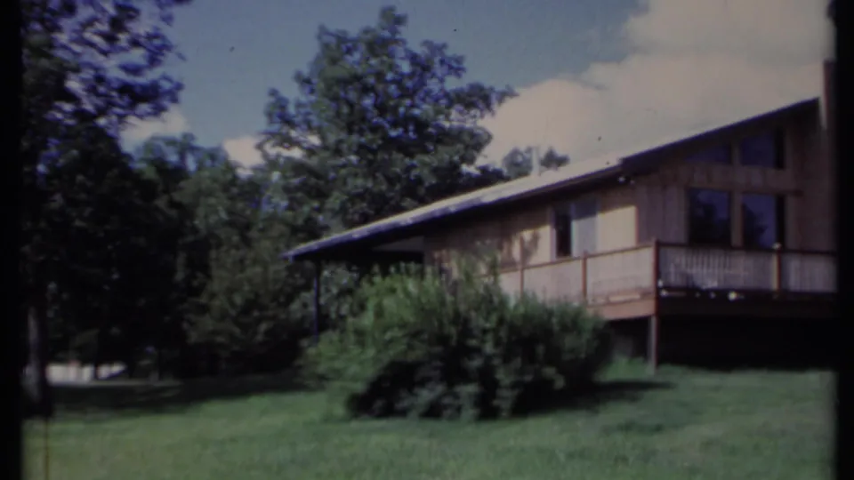 1971 California 8mm frame showing a side view of a light-colored home with a raised deck, lawn, shrubs, trees, and daylight sky.