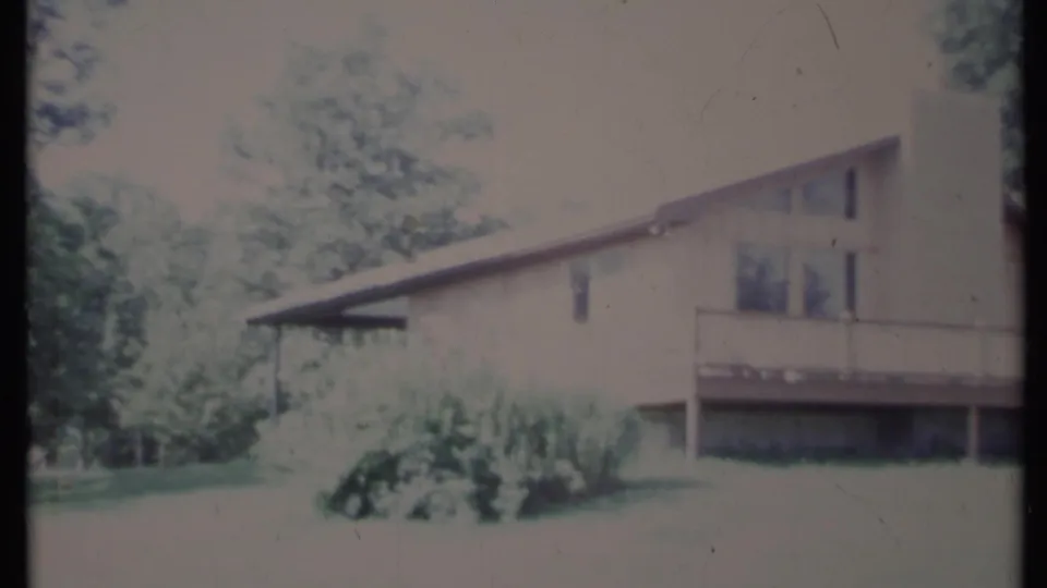 Soft 1971 California 8mm frame of a light-colored house side, raised deck railing, shrubs, trees, and lawn in bright daylight.