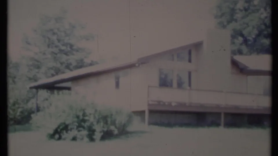 1971 California 8mm frame showing a light-colored home, long deck railing, large window, shrubs, trees, and lawn.