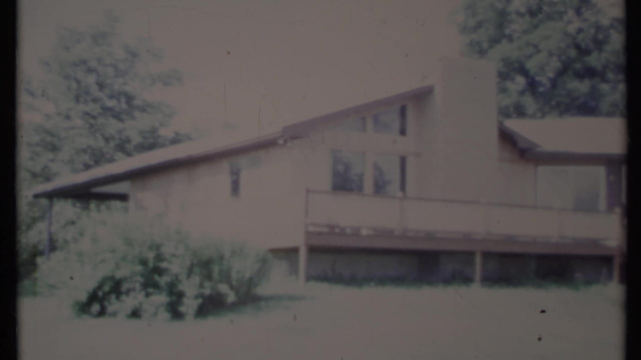 Soft 8mm frame of a sloped-roof house with deck, chimney, lawn, and trees.