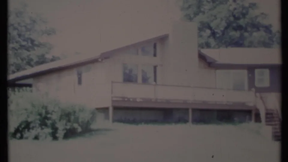 1971 California home-movie frame of a two-level house with upper deck, chimney, broad windows, lawn, and exterior stairs.