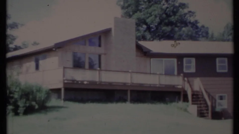 1971 California 8mm view of the house exterior, with deck posts, chimney, stairs, mixed siding, windows, lawn, and trees.