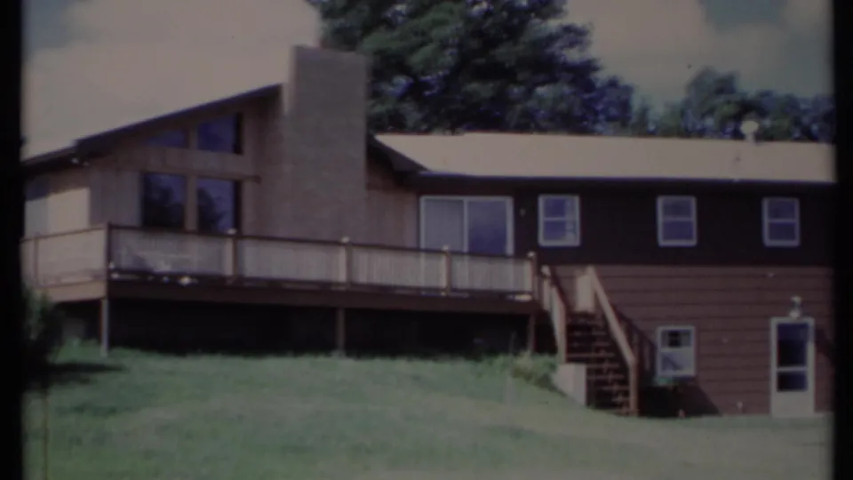 1971 California exterior frame showing the house's dark lower siding, upper deck, tall chimney, windows, stairs, lawn, and trees.