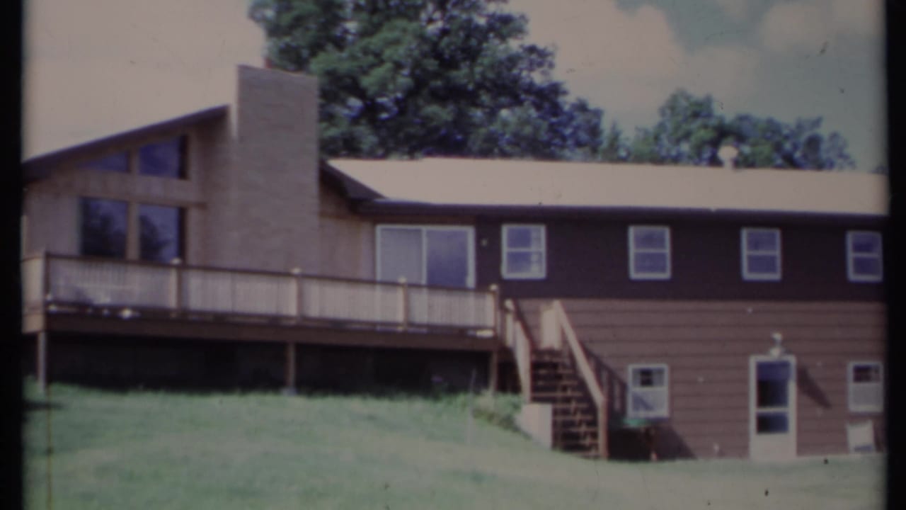 Brown two-story house with chimney, deck, stairs, lawn, and trees.
