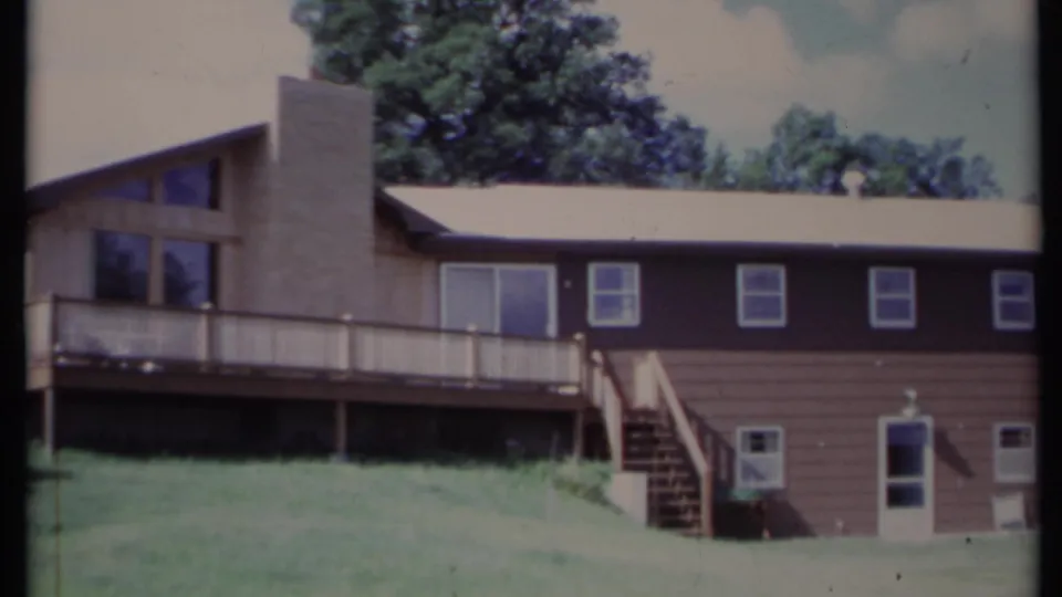 1971 California 8mm frame showing a brown two-story house with brick chimney, raised deck, exterior stairs, grass, and trees.