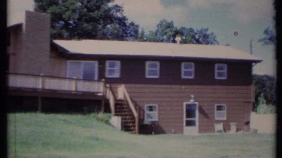 1971 California home-movie frame of the house exterior, showing the deck, stairs, upper windows, grassy yard, and trees.