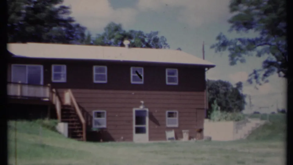 1971 California 8mm frame with the brown house facade, exterior stairs, raised deck edge, windows, lower door, and sloping lawn.