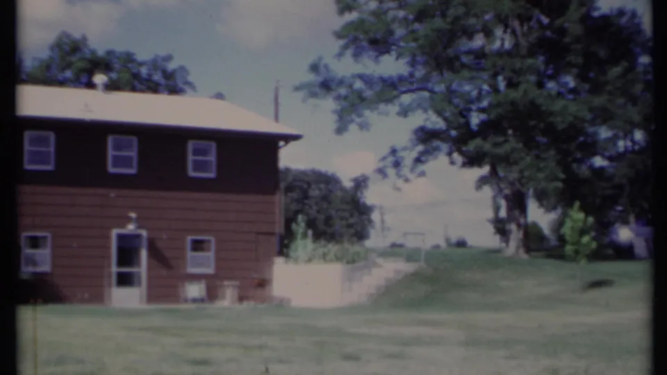 1971 California frame showing the right side of the brown house, lower doorway, upper windows, open lawn, large tree, and pale sky.