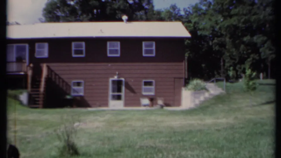 1971 California home-movie view of a brown house with deck stairs, lower door, lawn, and tree line.
