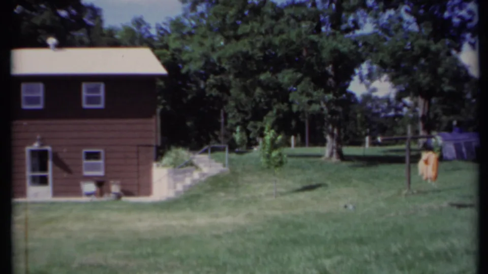 1971 California home-movie frame showing the brown house, open yard, trees, and a white caravan in the background.
