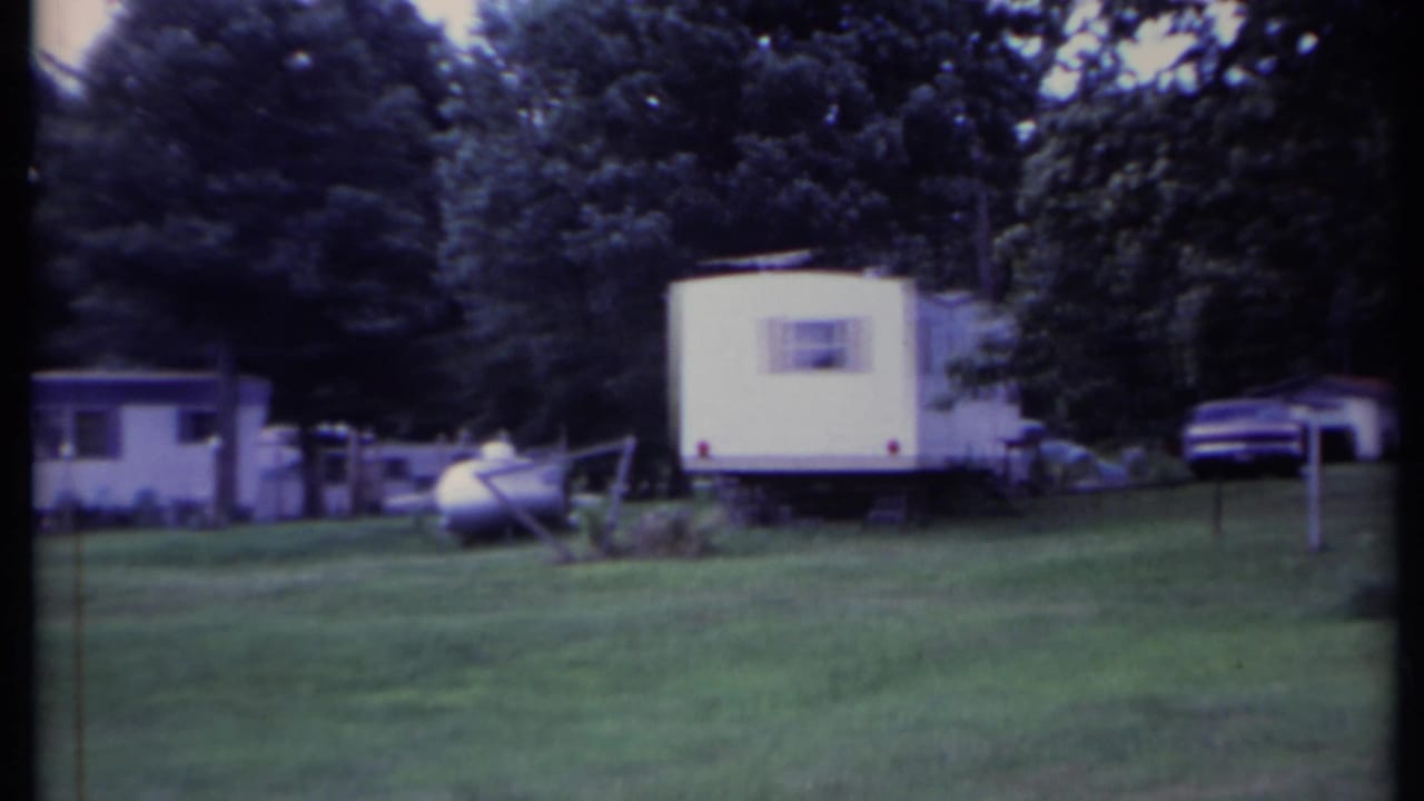 1971 California 8mm frame showing white mobile homes, lawn, trees, and a propane tank in a quiet park setting.