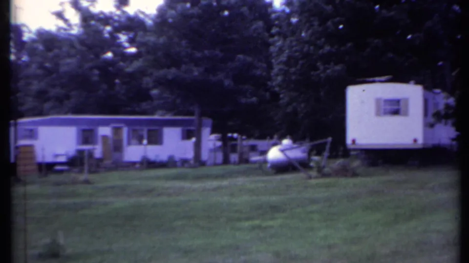 1971 California home-movie frame with a mobile home row, propane tank, lawn, and shaded trees.
