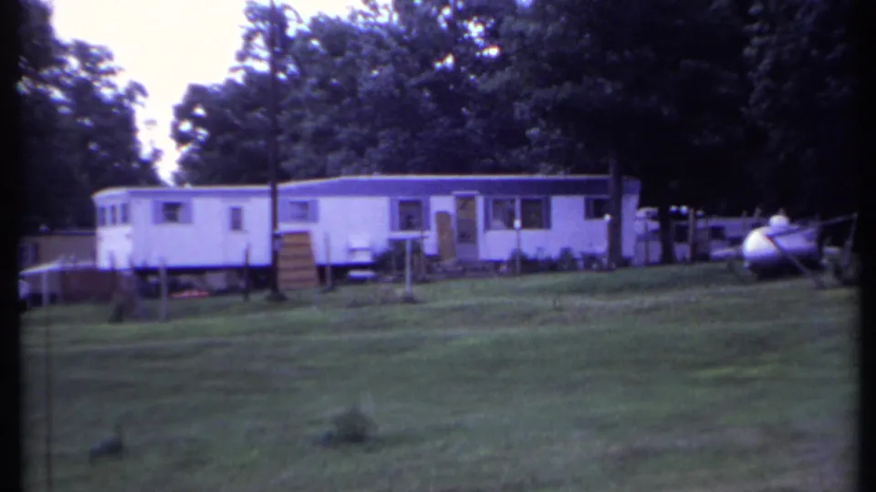 1971 California 8mm frame showing a wider mobile home park view with trailers, steps, posts, lawn, and propane tank.