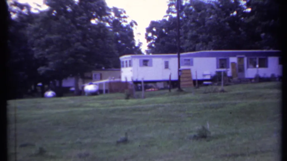 1971 California home-movie frame with multiple white mobile homes, open lawn, trees, yard posts, and a propane tank.