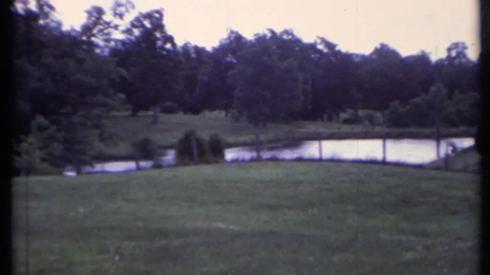 1971 California park view on 8mm film, showing open grass, still pond water, and wooded background.