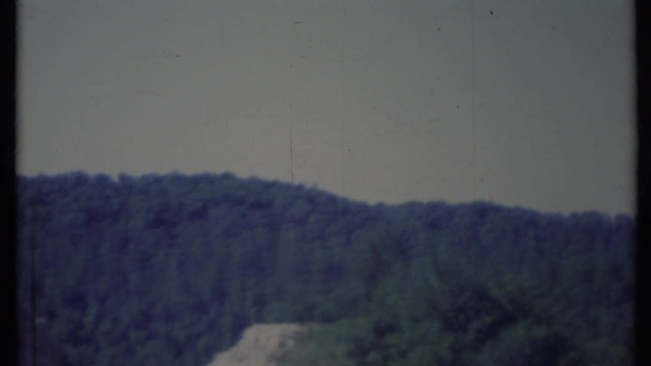 Dark wooded ridge with a pale road-like strip or clearing in the distance.