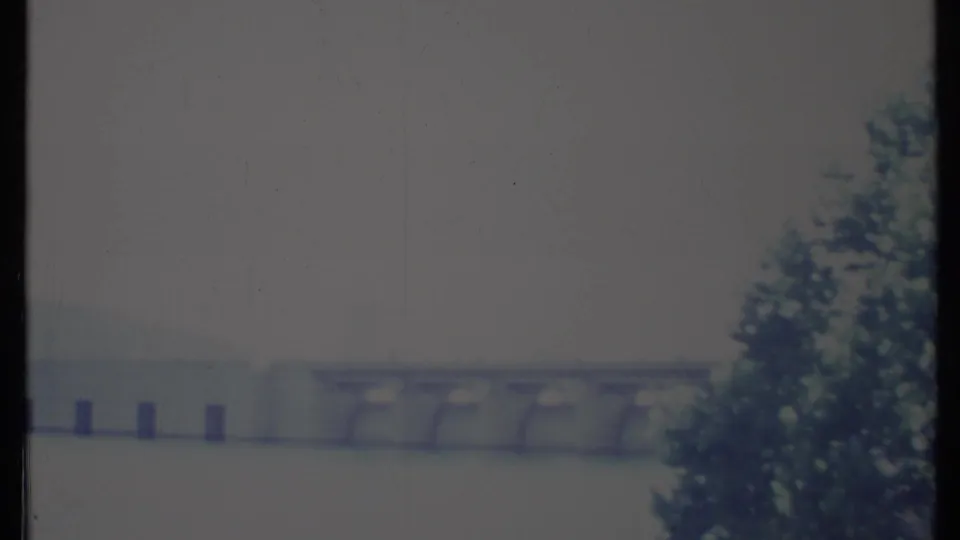 1971 California 8mm frame showing a concrete dam spanning water, with a hazy hillside behind it and foliage along the right edge.