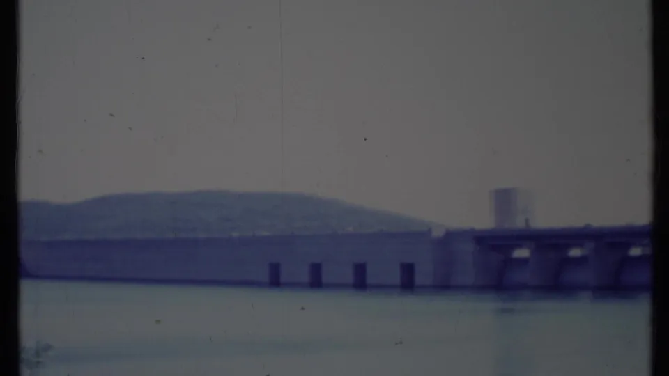 Final 8mm frame holds on the California dam face with vertical piers, still water, distant hill, and dark frame edge.