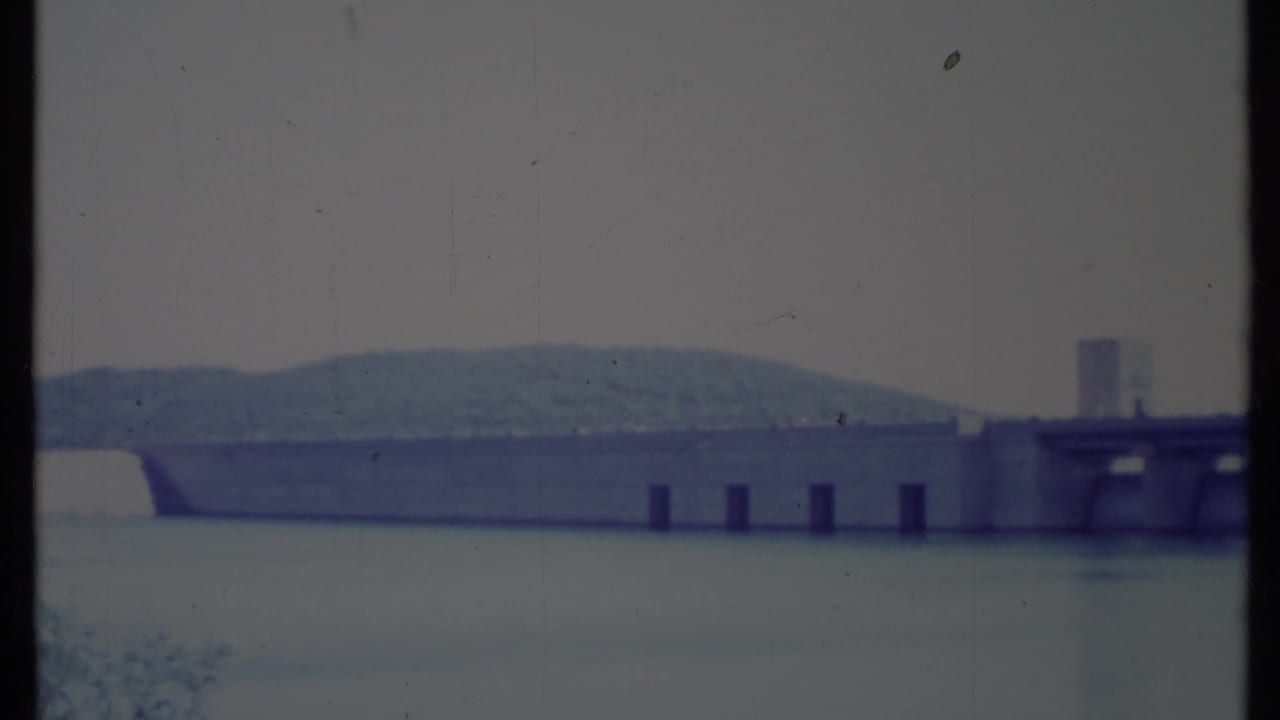 Wide view of a concrete dam across a reservoir with wooded hills behind it.