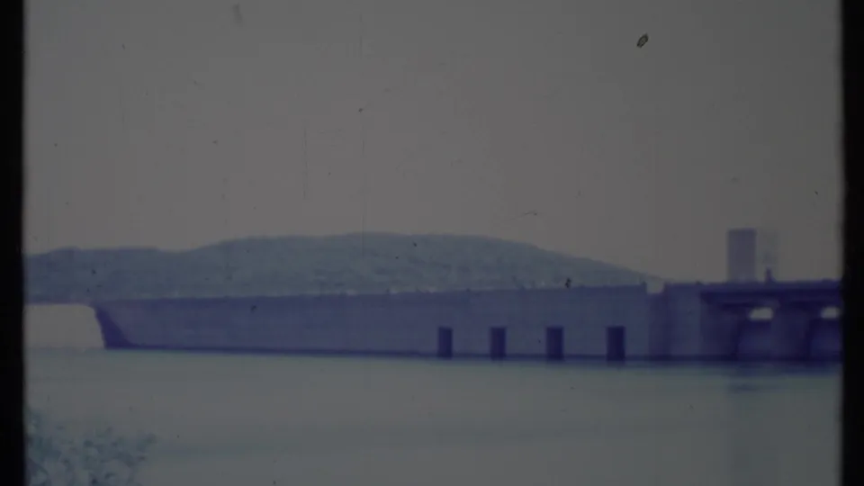 1971 California 8mm frame showing a long concrete dam across calm reservoir water, with low wooded hills and hazy sky in the background.