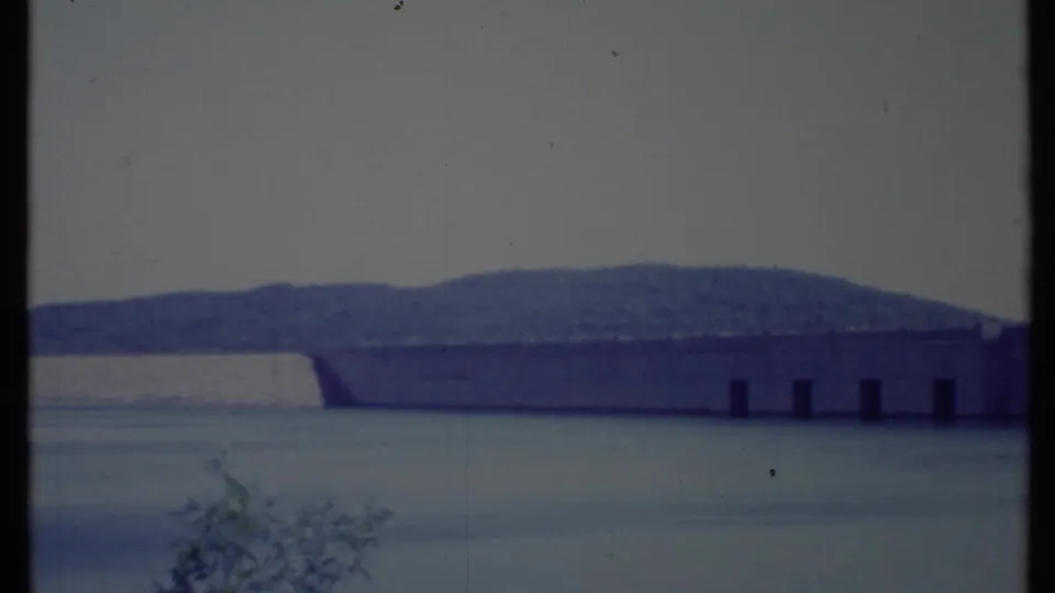 The dam extends across the reservoir in a soft 8mm view, with hills beyond and dark frame edges from the film scan.