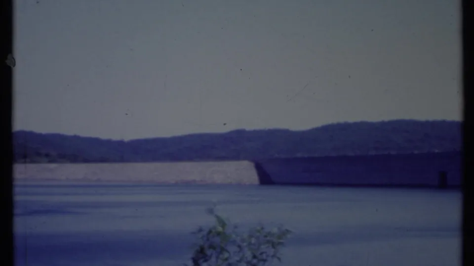 A wide 1971 California reservoir view shows the concrete dam wall, reflective water, distant tree-covered hills, and foreground brush.