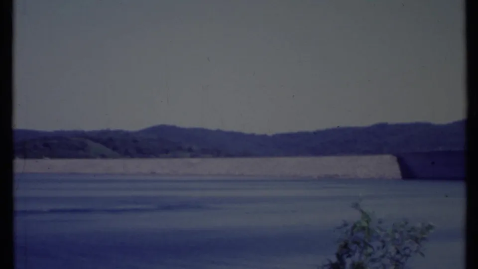 The closing frame shows the dam receding across a calm reservoir, with low California hills behind it and small brush at the lower edge.