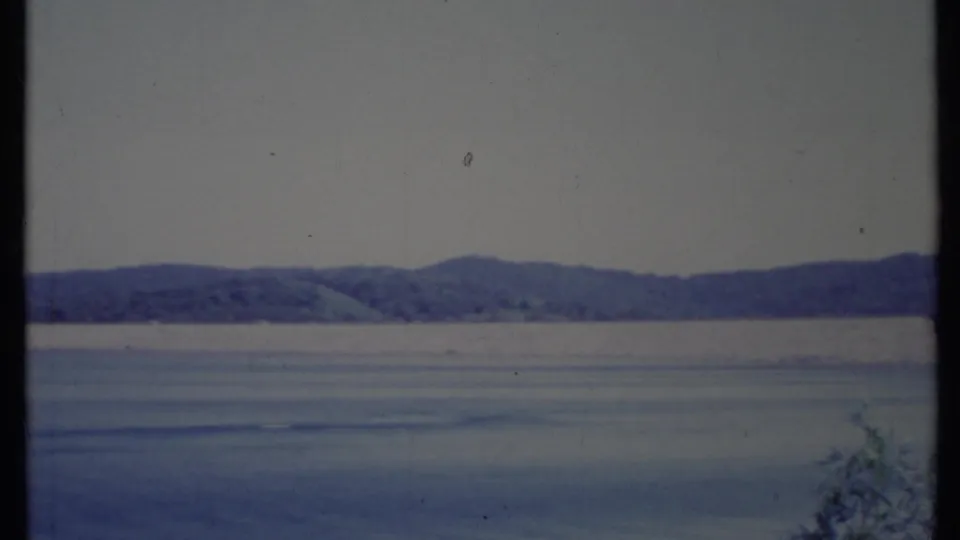 1971 California 8mm frame showing calm blue bay water, a distant tree-lined shore, and low hills beneath a hazy sky.