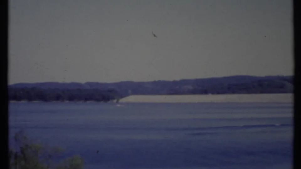 1971 California 8mm frame with gently rippled water, a narrow far shore, low hills, and faint foreground vegetation at the lower edge.