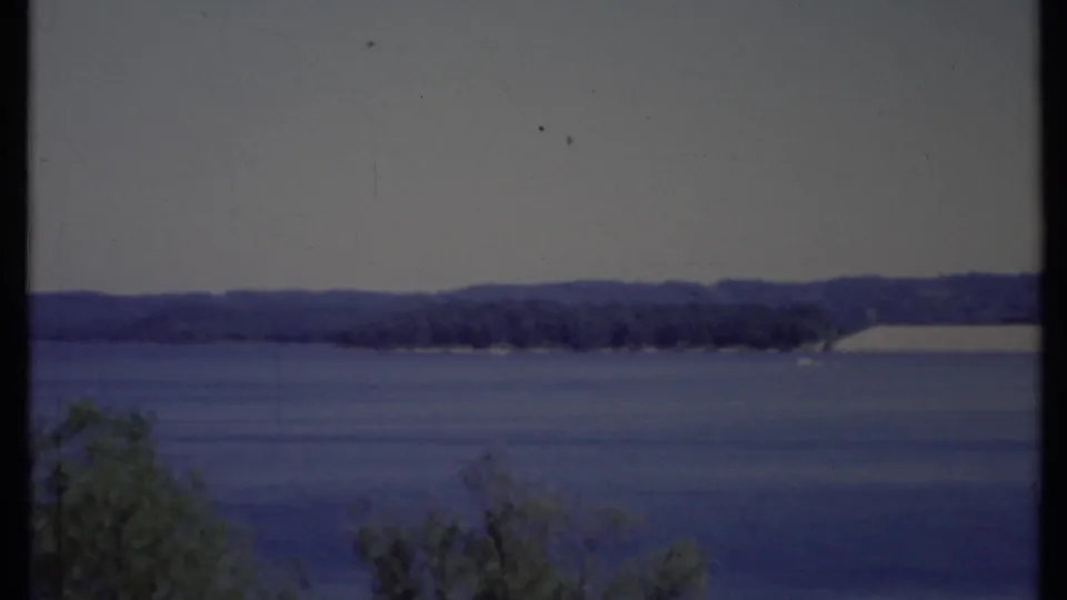 1971 California 8mm frame looking over blue water to a distant shore, with dark gate edge and shrubs visible in the foreground.