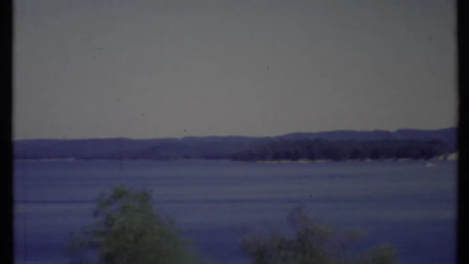 1971 California 8mm frame showing a broad blue lake, distant low hills, pale sky, and soft foreground trees.