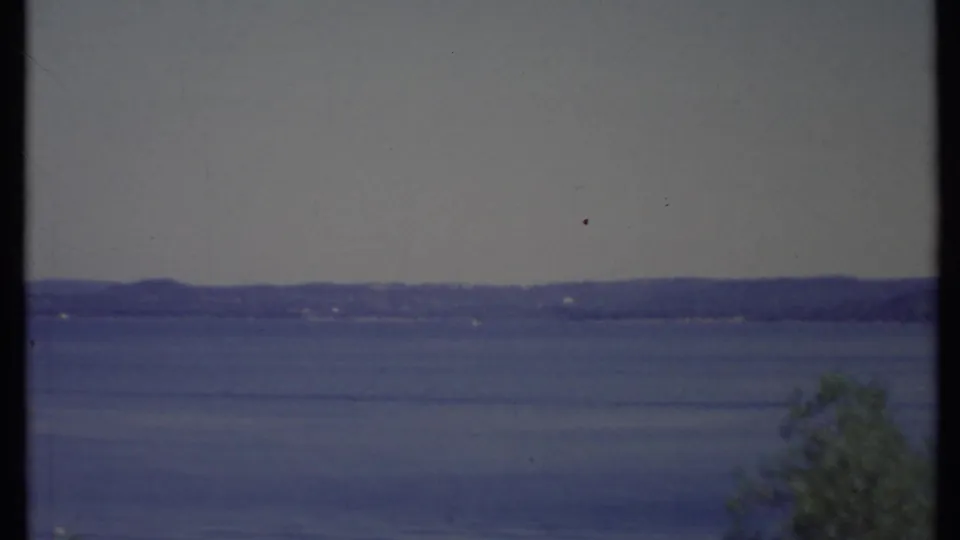 1971 California 8mm view across a blue lake toward low hills, with a small foreground tree and faded film color.