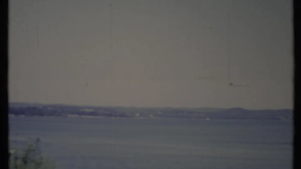 Faded 1971 California 8mm frame of a hazy lake, low distant hills, pale sky, and sparse foreground tree tops.