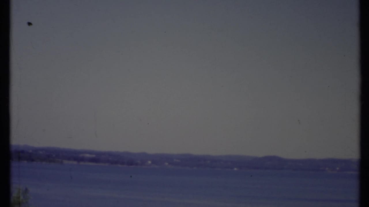 Distant shoreline across blue water under a muted sky, framed by dark vertical film edges.