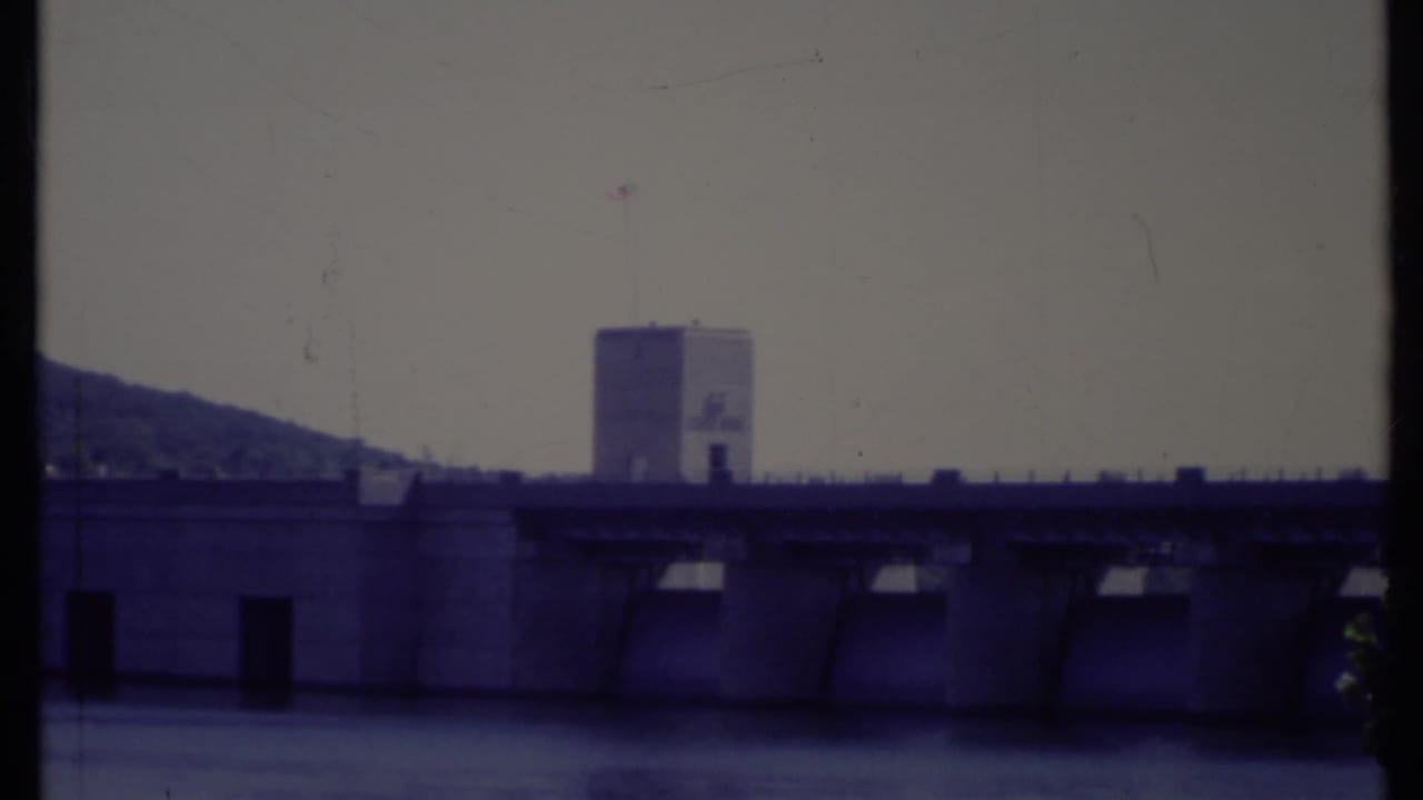 Dam with spillway bays, tower, flag, hillside, and water in soft focus.