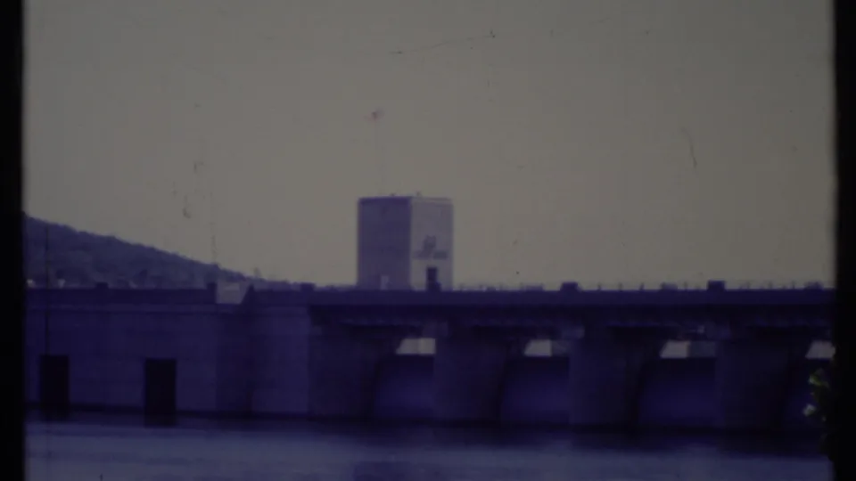 1971 California 8mm frame showing a large dam, tower with American flag, water, and a wooded hillside beyond.