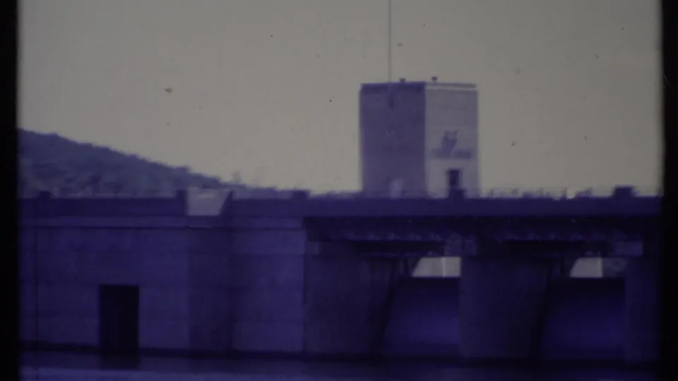 1971 California 8mm frame showing the dam tower with flag, dark doorway, spillway bays, and water in the foreground.