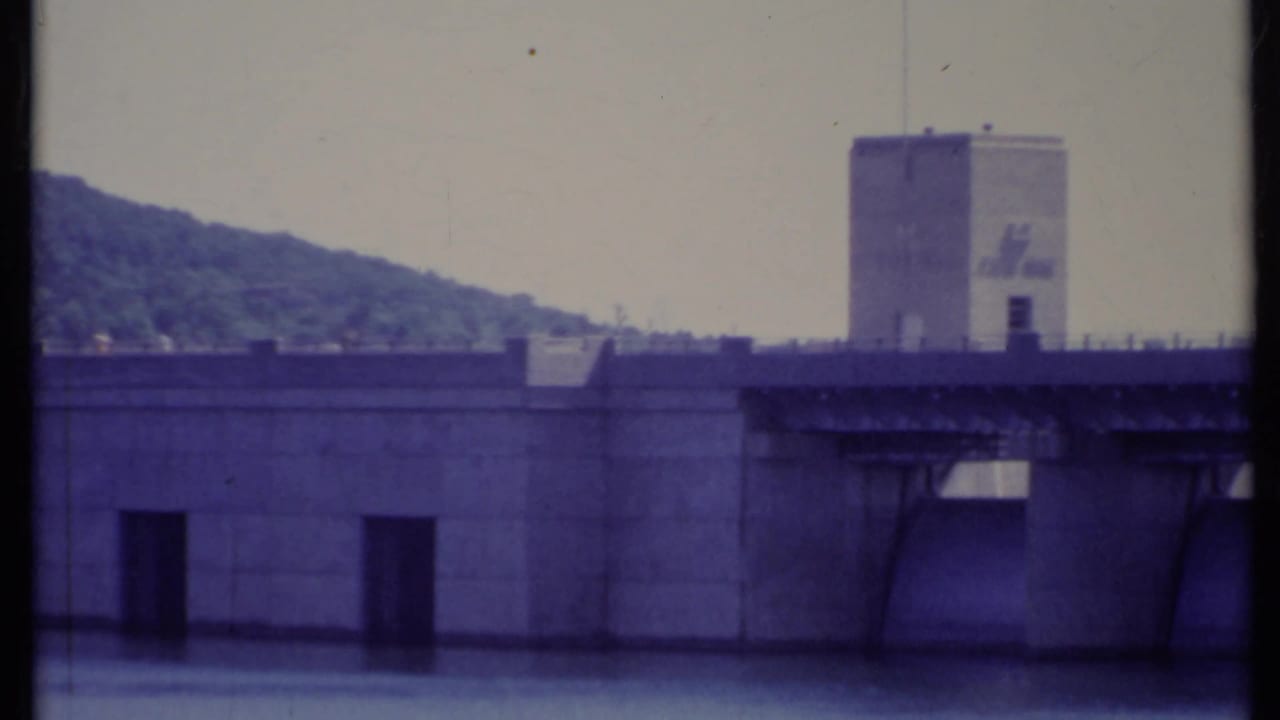 Concrete dam with tower and wooded hillside behind it.