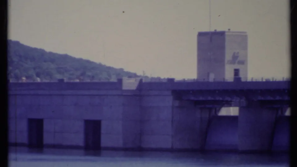 1971 California 8mm frame showing a broad concrete dam, still water, a tower, and a forested hill in the background.
