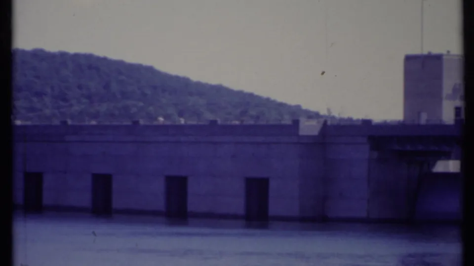 1971 California home-movie frame of a concrete dam wall with multiple dark openings and a tree-covered hill beyond.